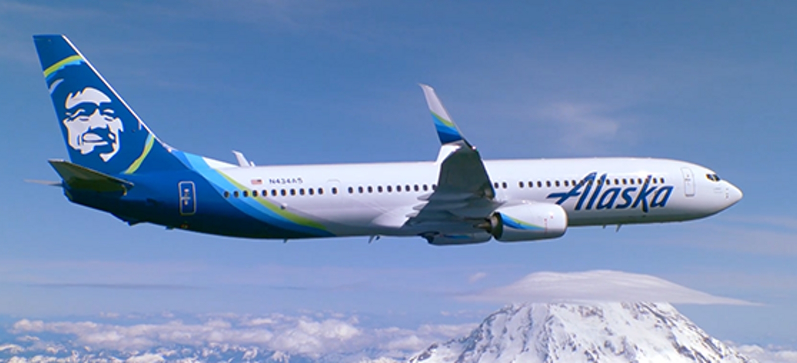 An Alaska Airlines jet flies over the Cascade Mountains in a clear sky.