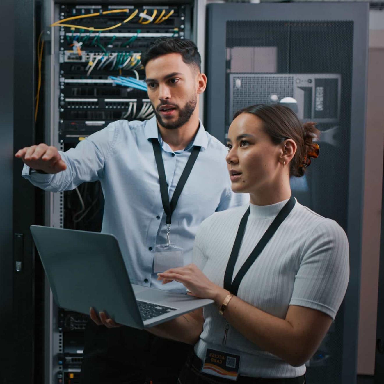 An IT professional holds a laptop and engages in a conversation with a colleague in a server room.